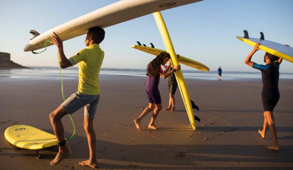 Children balance boards on their heads as they walk to the shore's edge.