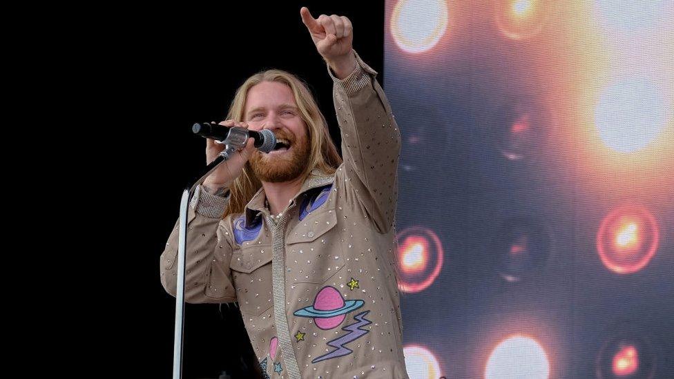 Sam Ryder pointing to the crowd as he holds a microphone in his other hand and smiles and sings