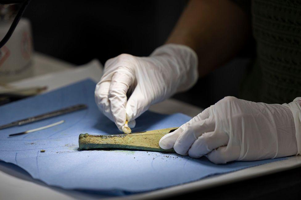 A pair of white latex-gloved hands work away delicately at a Bronze Age item covered in dirt and rust. It is on a sheet of blue paper with other conservation tools sitting to one side