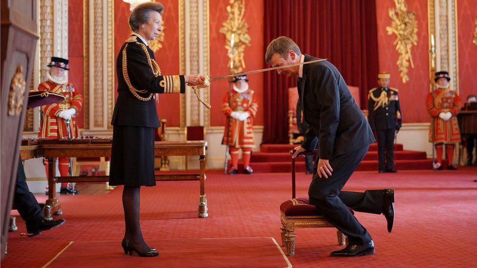 Ian Rankin kneels on a stool as he is knighted with a sword by Princess Anne  in a palatial room with a red carpet and red and gold walls. There are royal  members of staff in the background.