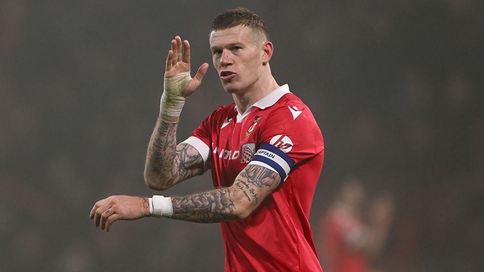 James McClean wearing the red football shirt of Wrexham during a match. He has short brown hair and and tattoos on both arms he is wearing the captains armband and has his right arm raised which is bandaged