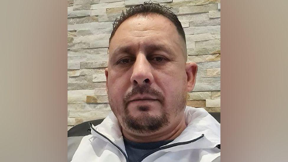 Robert Calin, a man with short dark hair and a goatee beard, wearing a grey and white tracksuit top and pictured in close-up against a brick wall