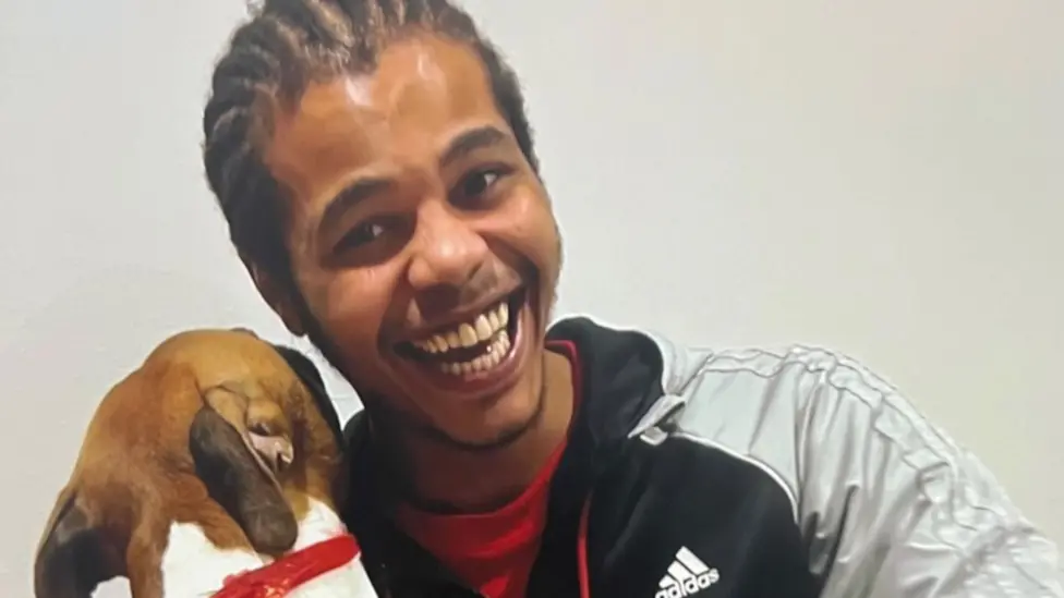 Kyron Lee, who is wearing an Adidas sports jacket that is silver and black, a red T-shirt, has dreadlocked black hair, and is smiling at the camera while hugging a dog, which has its back to the camera 