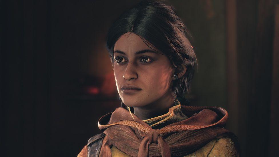 A screenshot of a young woman in a dimly lit room. She's got loosely tied back hair. She's wearing a ragged, hooded garment with signs of wear and tear from frequent adventuring.