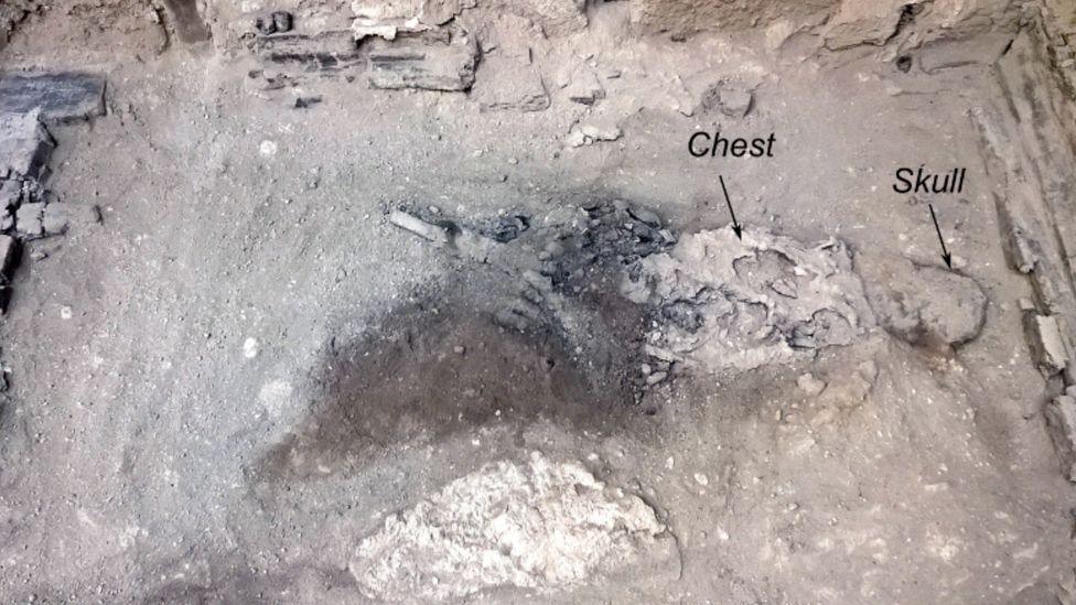 A picture of the remains of the man. The remains lie on what looks like greyish sand - a skull, more or less the same colour as the sand, a chest-thorax that is whiter in colour and the bottom part of the skeleton, which is of a material darker in colour, almost looking like charcoal. The area is surrounded by greyish brick walls.