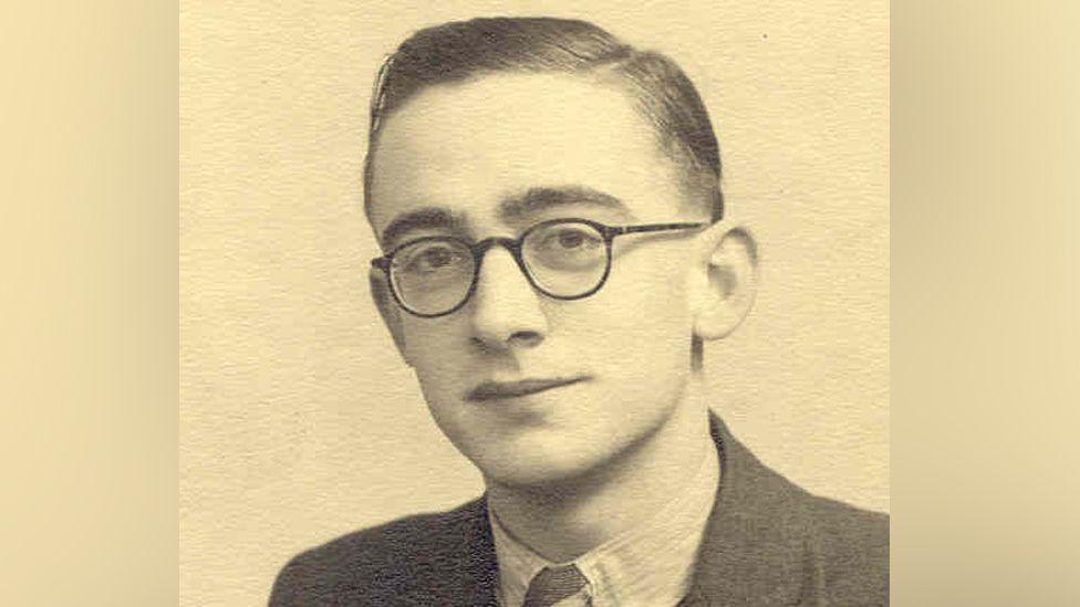 Black and white photo of John Teague with short hair swept to the side and round glasses. He is smiling at the camera. He is wearing a shirt, tie and jacket.