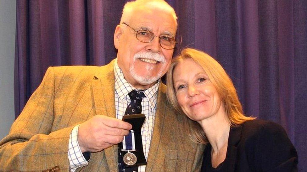 A full-colour picture of a man showing a military medal as a woman leans on his left shoulder