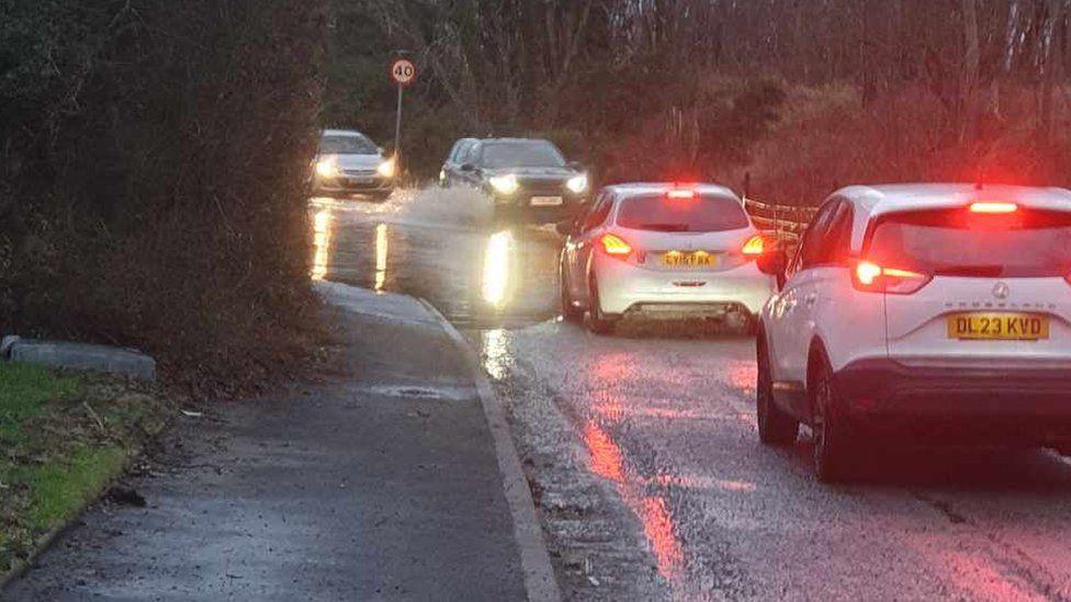 Two cars on the left hand side of Gobowen Road with their red brake lights on as they drive through standing water, which stretches across the width of the road. There are two cars coming in the opposite direction on the right hand side of the road. They have their headlights on. The front car is driving through the water, and there is water splashing up around the front wheel.