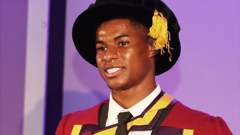 Marcus Rashford wearing a black cap with a yellow tassel and a red university gown.