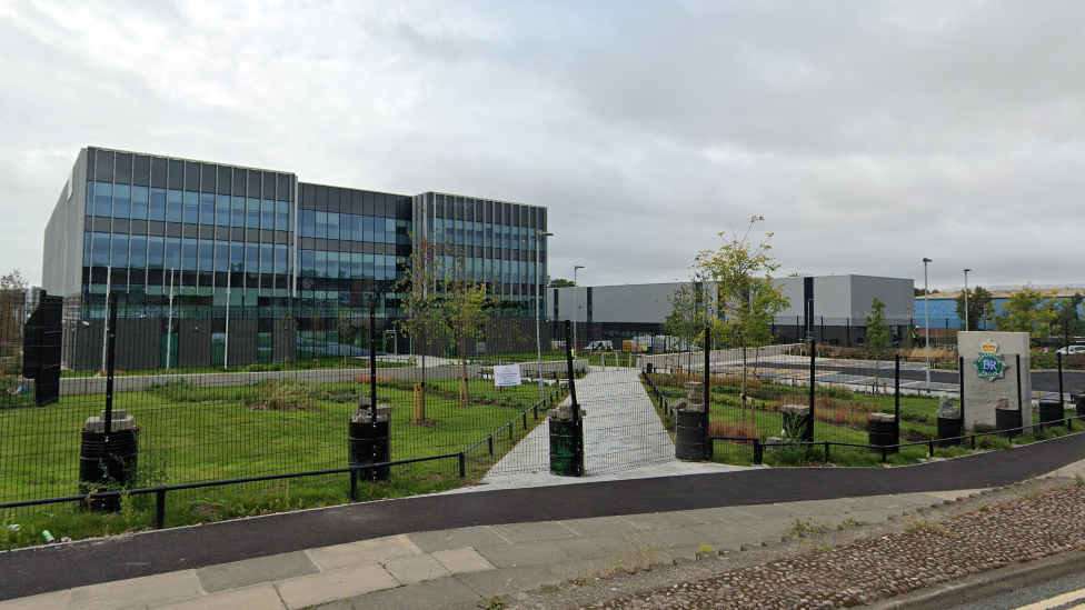 Merseyside Police HQ, a large rectangular glass fronted building with grey cladding, and a grassy area and car-park behind a fence in the foreground.