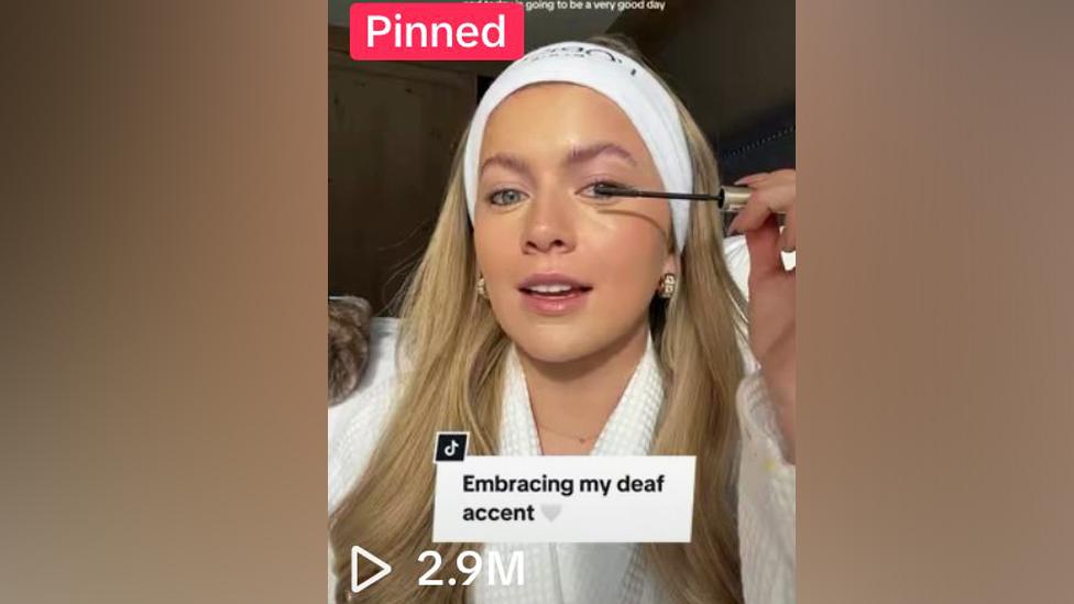 TikTok video showing Tasha, a white woman, holding a black mascara wand, while applying it on her left eyelash. She has a white headband on and is wearing a white gown. The title of the video written in a white box and black font is "embracing my deaf accent" white a white heart emoji. Under that is a white "play" sign with 2.9m written. At the top of the screenshot is a "pinned" caption written in white on a pink box.