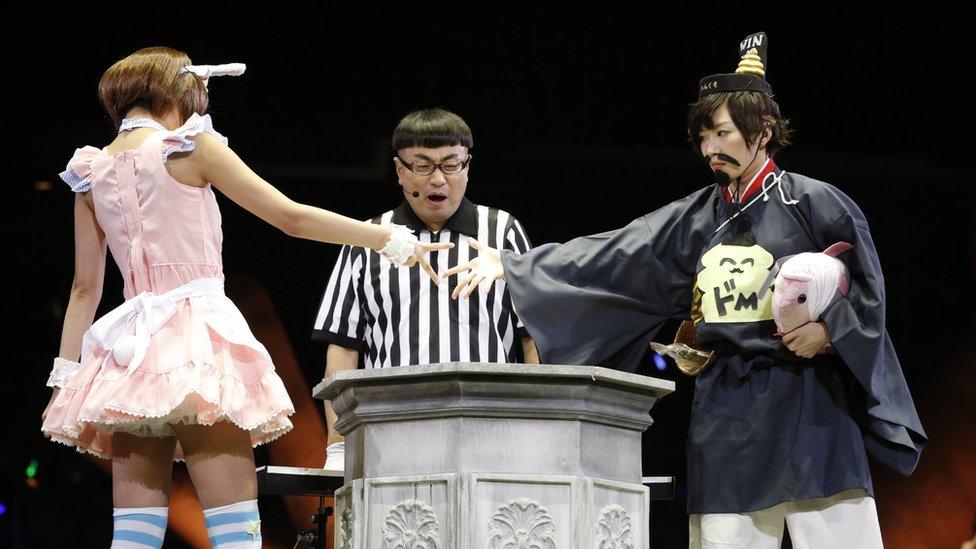 Two women compete in the contest in 2014, with the woman on the left, in a pink dress, throwing "scissors", to defeat the woman on the right, wearing a fake moustache and dressed in a bizarre re-imagining of traditional Japanese clothing.