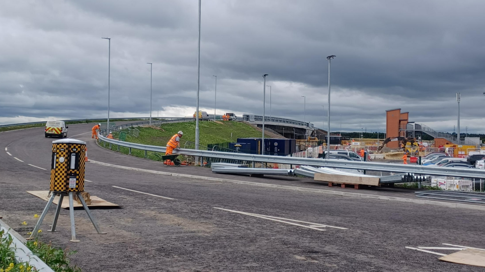 A large modern road bridge sweeps over a new railway station with construction workers wearing orange hi-vis jackets and trousers working on the barriers. Under the bridge construction work with diggers continues 