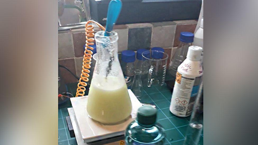 Chemicals in glass containers and bottles on a desk. The desk is green and several chemical bottles can be seen. Multi-coloured tiles are behind.