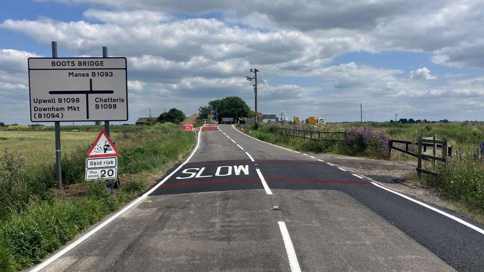 A road with slow written in large white letters on black tarmac, with red countdown markers, and a sign saying Boots Bridge on the left