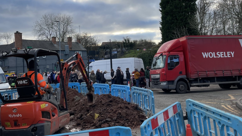 A red digger next to a pile of rubble, surrounded by light blue plastic fencing. A queue of people can be seen in the background