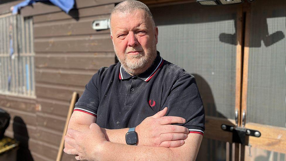 Pigeon fancier Paul Smith, standing in front of a pigeon loft on his allotment, wearing a dusty polo shirt, with his arms crossed.