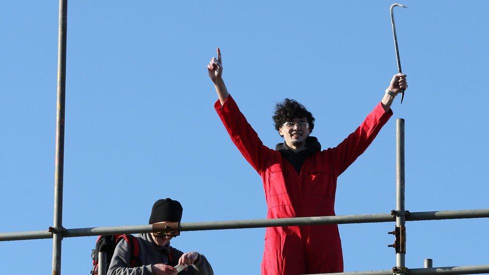 A man with dark curly hair wearing a red overall holds his hands aloft while holding a metal crowbar