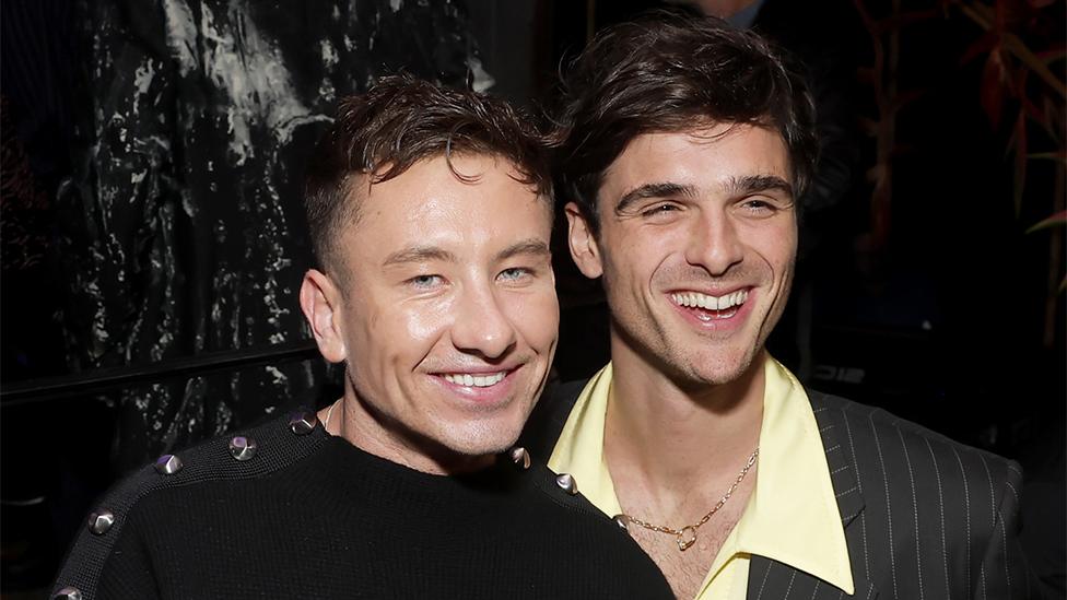 Barry Keoghan and Jacob Elordi smiling, with arms around each other on a black background. Barry is wearing black with jacob wearing a yellow shirt and black striped blazer.