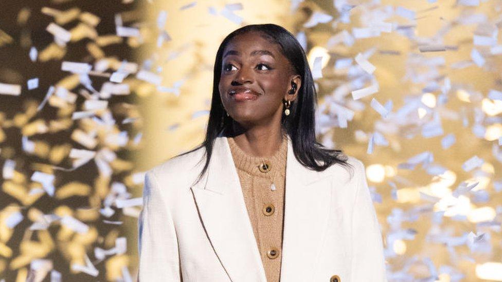 Cat on stage, she's wearing a white suit over a beige jumper, the stage backdrop is gold and confetti falls around her.