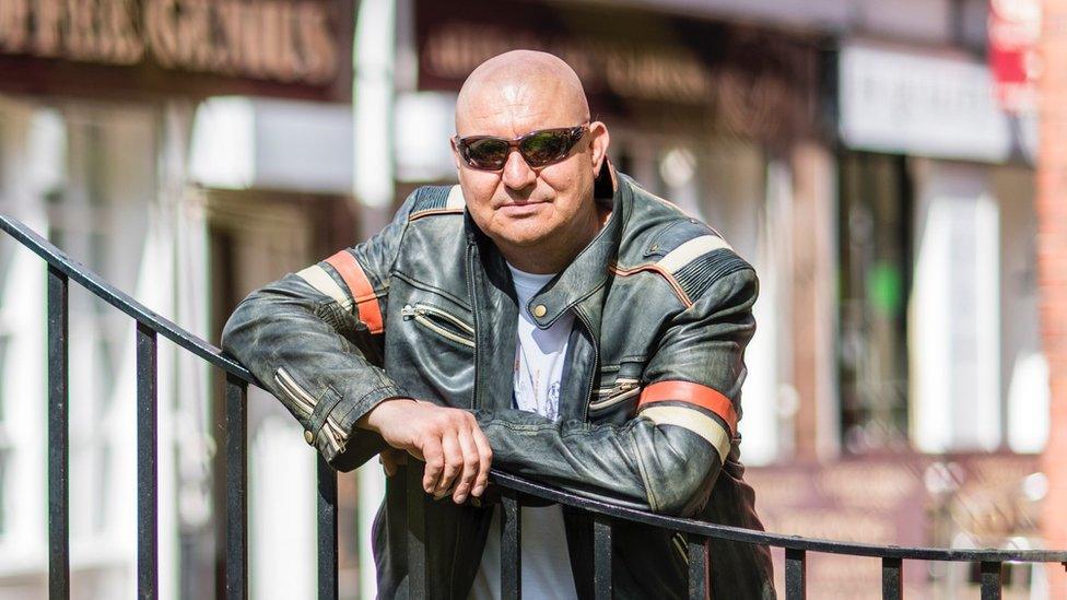 Mike Craven wearing sunglasses and a leather jacket leans on a metal fence