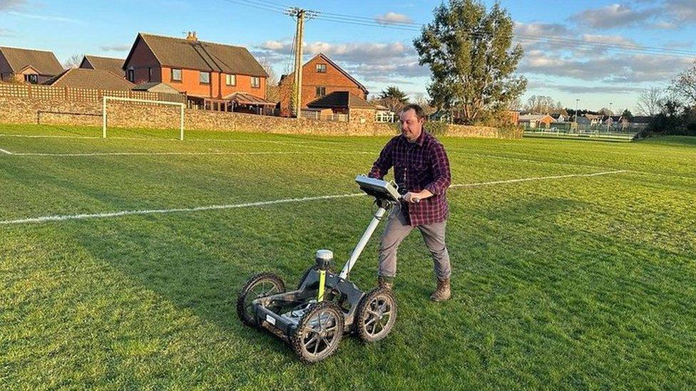 Dr Henry Webber walking along a playing field pushing a four-wheeled contraption