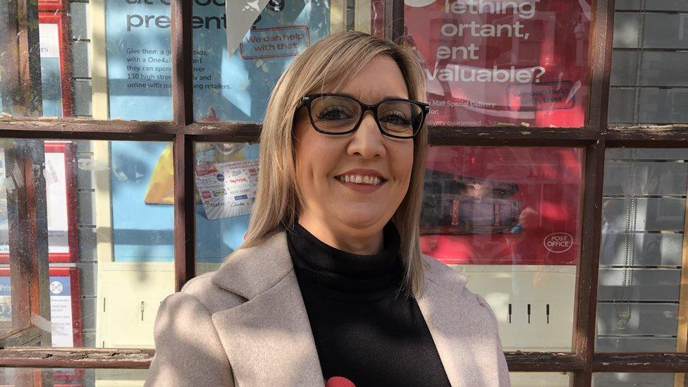 Melissa Whittaker looks into the camera. Wearing glasses, a grey jacket and black top. Stands in front of a shop window.