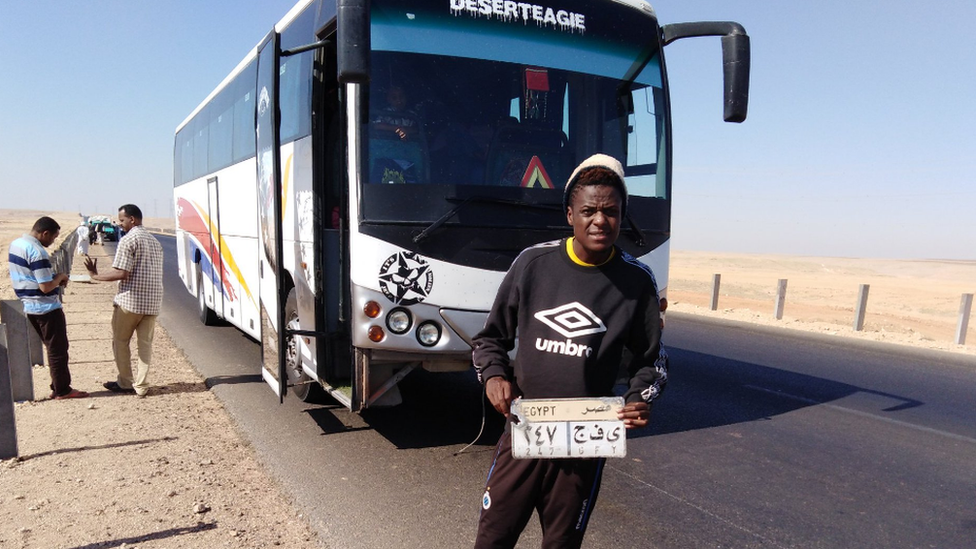 Alvin Zhakata holding up the licence plate of a coach in the desert en route from Sudan to Egypt