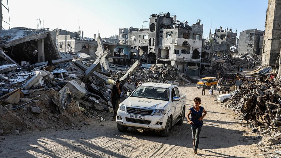 A boy in dark trousers and a sleeveless shirt is running up a street in Gaza passed a white truck marked UN. He is surrounded by the wreckage of buildings with a badly damaged but still standing building in the background. A yellow people carrier can be seen at the other end of the road.