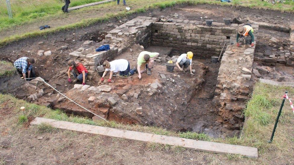 Six people use hand trowels to dig around old stone walls