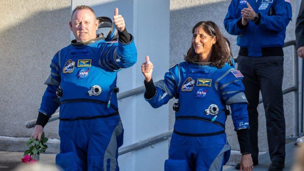 Astronauts Butch Wilmore and Suni Williams getting ready to travel to the International Space Station. They are wearing blue suits and are giving a thumbs up signal.