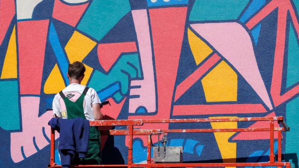 A man standing on a red mechanic lift painting a large mural on a wall