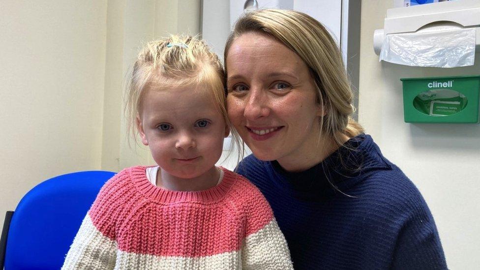 Elinor and her mother, Victoria. They are sat in a doctor's evaluation room on a blue chair. Elinor is a young, blonde child. She is wearing a pink and cream jumper. Victoria is sat next to her, with her cheek on hers. She is wearing a navy blue jumper. They are both looking directly at the camera. Elinor has a blank expression and Victoria is smiling.