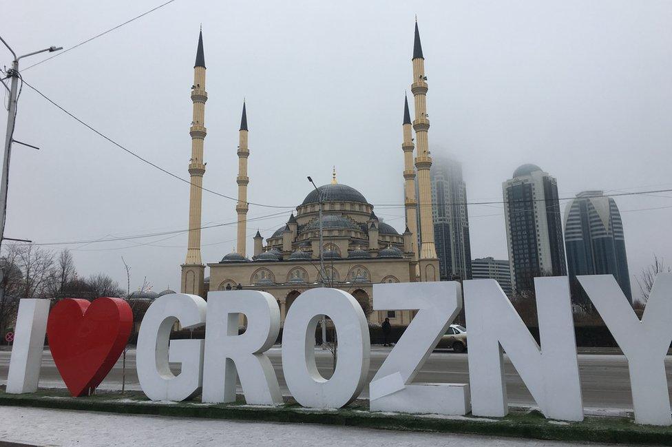 A large street sign reads: "I (heart symbol) Grozny, in front of a church, with skyscrapers seen in the background