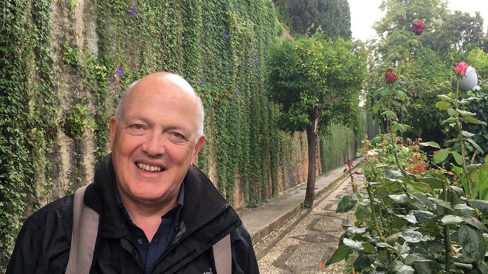 Klaus Vella Bardon is pictured in a manicured garden, with lush greenery stretching along the path behind him