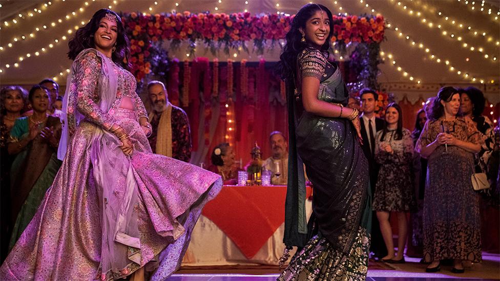 Characters of Kamala and Devi dancing. Kamala is standing on the left wearing a shiny purple outfit. Devi is on the right wearing a glittery black outfit. The background is full of lights and flowers on the walls and ceilings, with many people standing behind the characters watching the dance