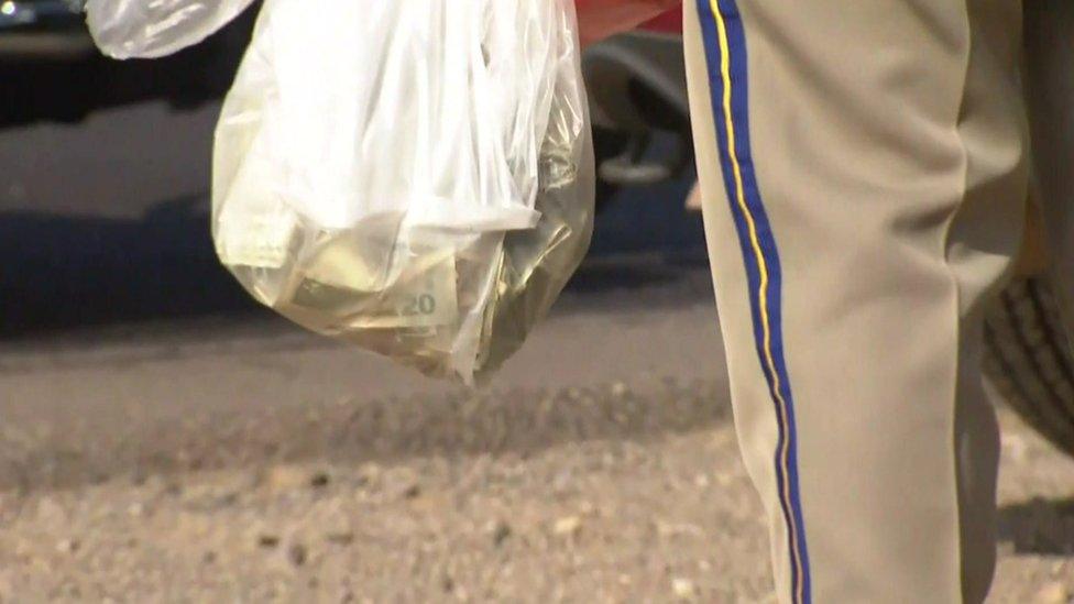 A police officer holds a carrier bag filled full of US dollars