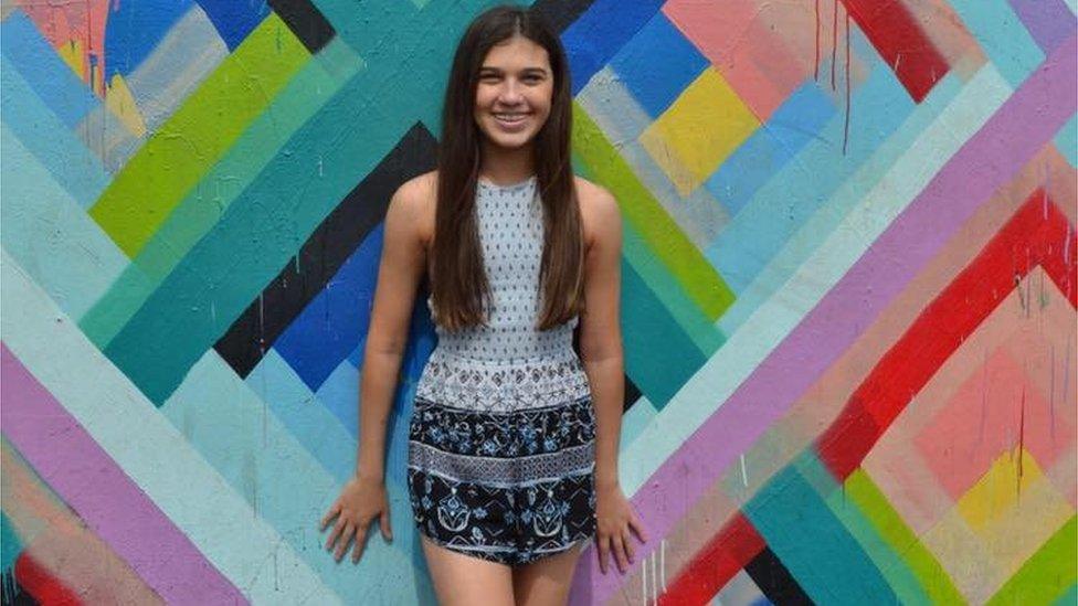 A teenage girl stands against a bright wall and smiles