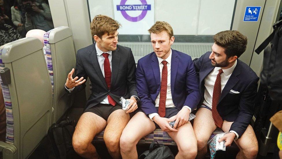 Three trouser-less men in suits sit and chat on the Elizabeth Line at Bond Street station.