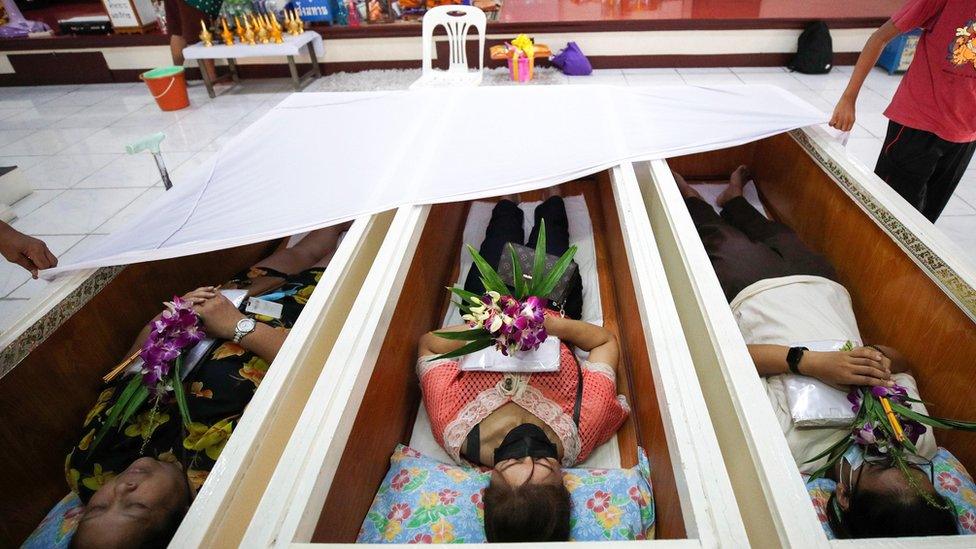 Thai people lie down inside coffins as they offer prayers with flowers, candles and joss sticks