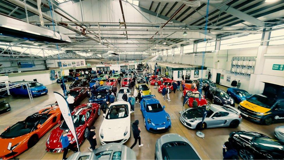 An indoor hanger full of cars with people around the edge studying them