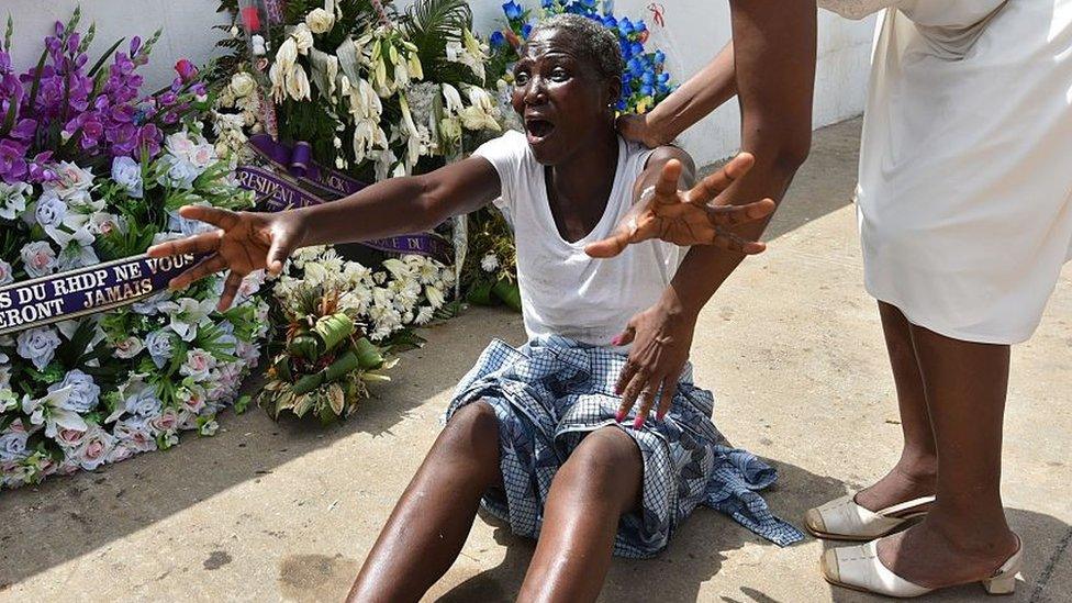 A woman, helped by a relative, cries next to wreaths of flowers in front of the Etoile du Sud hotel on March 20, 2016 in Grand Bassam, during a tribute to the victims of a djihadist attack that killed 19 people on March 13