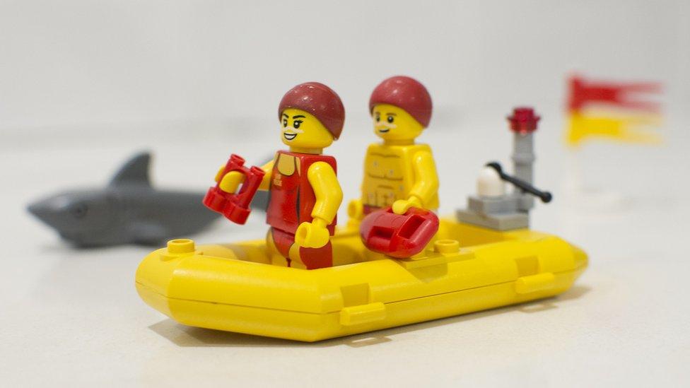 Lego lifeguards on patrol in an inflatable rescue boat. A shark lurks in the background. A red and yellow flag flies in the breeze.