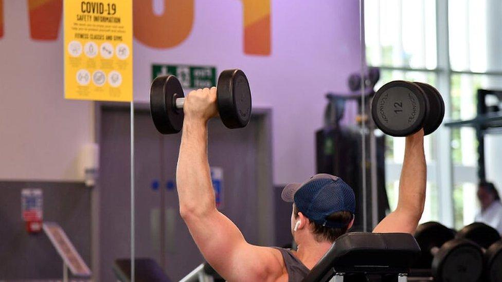 A man lifting weights in a gym in front of a coronavirus safety sign