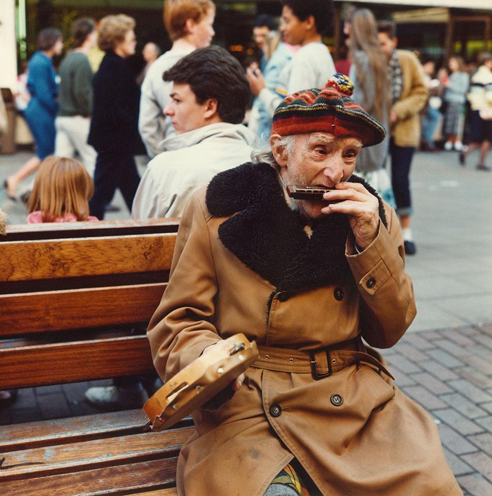 An elderly man sits on a street bench and plays the harmonica