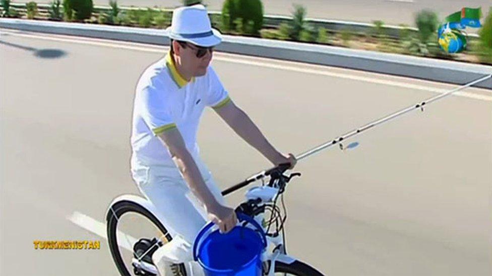 Turkmen president riding a bike, with a fishing rod in one hand, a bucket in the other