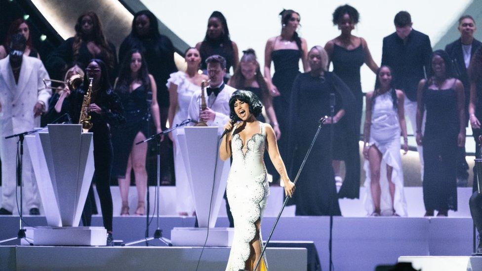 Raye performing at the 2024 Brits with choir Flame Collective behind her. Raye has a short dark bob and wears a long white dress with black crystal detailing and scalloped edges. Behind her are musicians with brass instruments as well as the singers from the Flame collective choir. The other musicians are all dressed in black or white