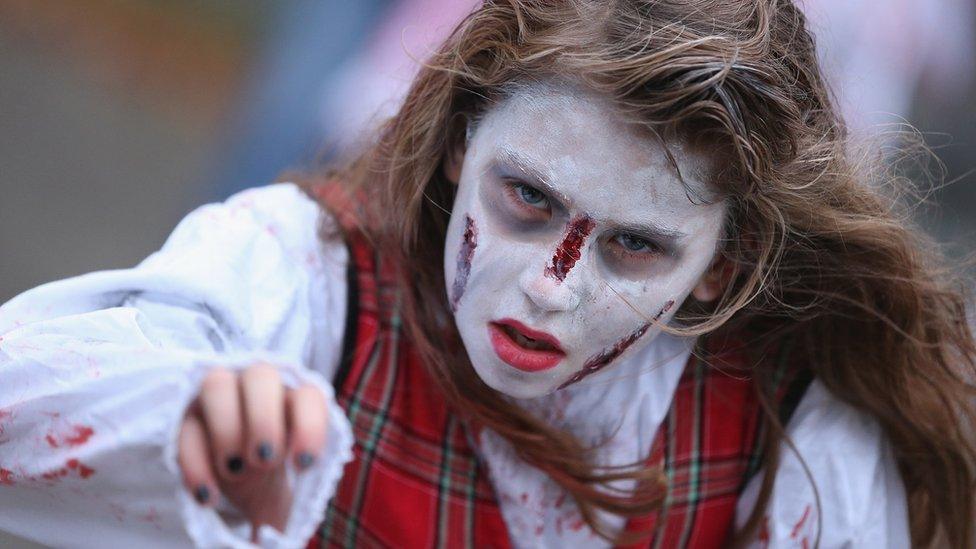 A woman with her face caked in white and with red painted scratches on her face stares lifelessly reaching forward, imitating a zombie.