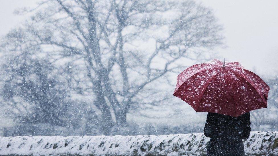 A woman wearing a long winter coat shelter under a read umbrella in a snow storm next to a snow-covered stone wall.