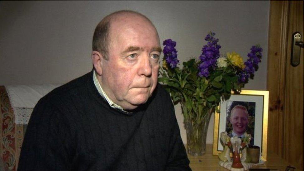 Colin Bell in a black jumper, pictured in front of a framed photo of his son Kevin and purple flowers in a vase